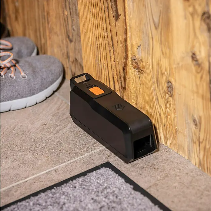 Windhager Mausefalle elektronisch - Anwendungsfertig, Schwarz - Eine elektronische Rattenfalle in Schwarz mit einem orangefarbenen Knopf steht auf einem Boden neben einem Schuh.