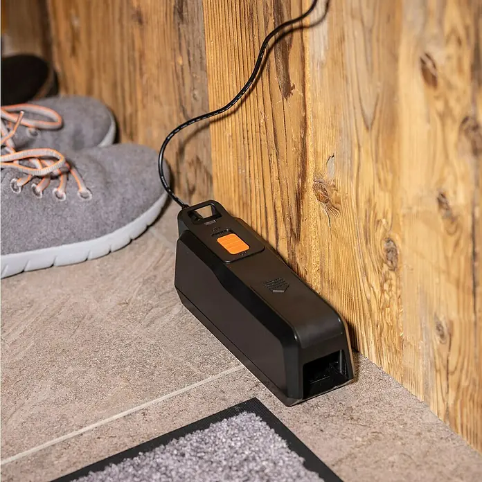 Windhager Mausefalle elektronisch - Anwendungsfertig, Schwarz - Eine elektronische Rattenfalle mit einem orangefarbenen Knopf, die an eine Holzwand gelehnt ist, dient zur effektiven Nagetierbekämpfung.