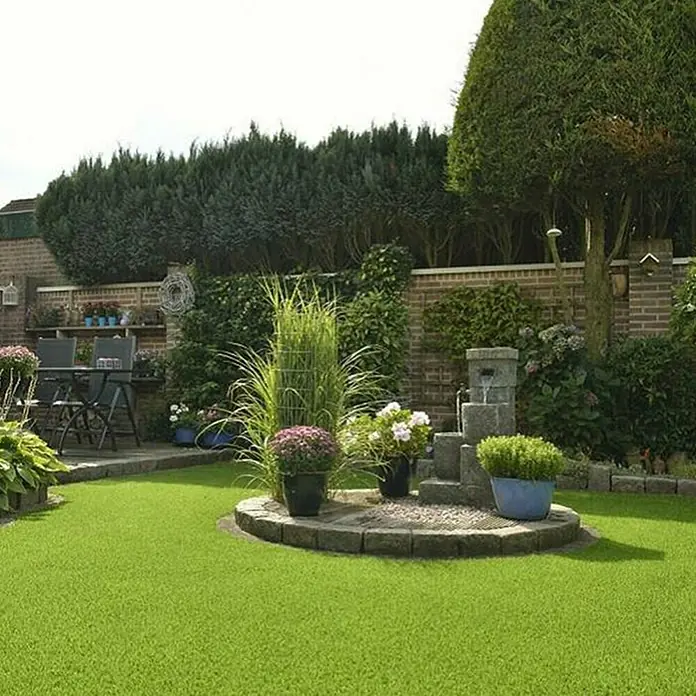 Kunstrasen Oxford - 1.900 x 200 cm, Grün - Ein grüner Garten mit einem Springbrunnen, Pflanzen und einem gepflegten Rasen, der eine friedliche und ästhetisch ansprechende Atmosphäre schafft.