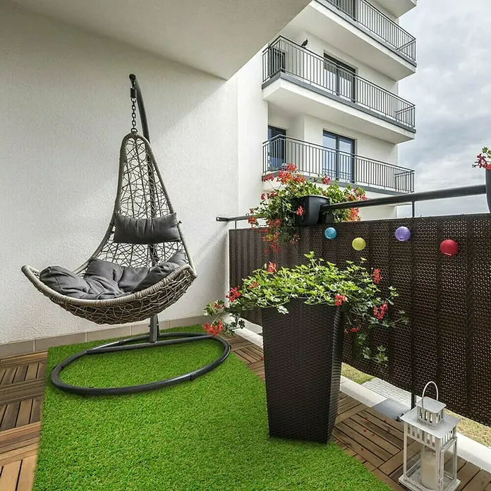 Kunstrasen Riviera - 400 x 200 cm, Grün - Einladende Balkonecke mit einem hängenden Rattansessel mit weichen Kissen, grüner Kunstrasenfläche und blühenden Blumenkästen.