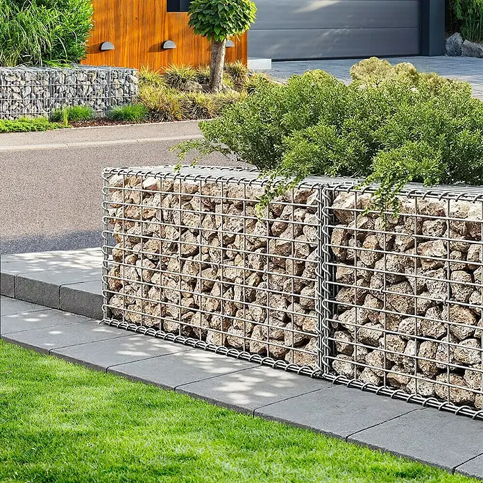 Floordirekt Gabione Lenox - 30 x 100 x 50 cm, Stahl, Maschenbreite: 10 cm, Maschenhöhe: 5 cm - Bild einer Gabionenwand im Garten, die mit Steinen gefüllt ist, mit Rasen und Pflanzen im Hintergrund.