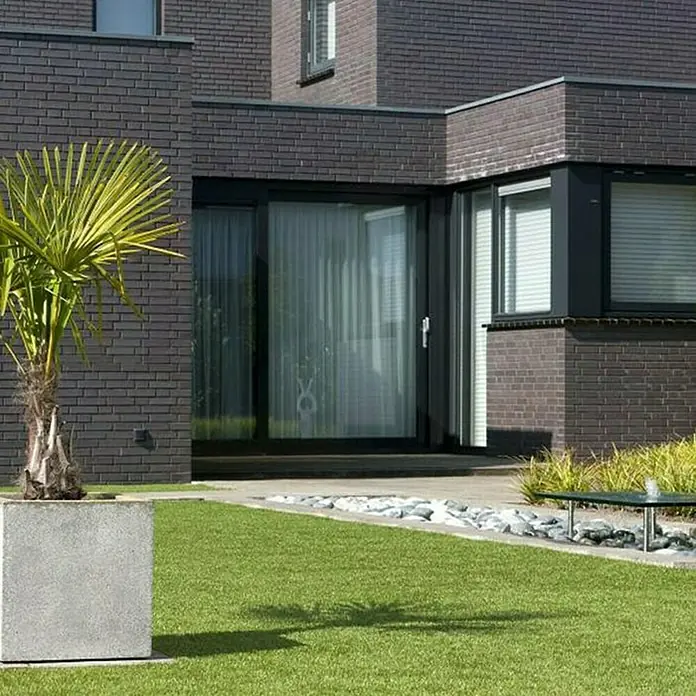 Kunstrasen Windsor - 100 x 100 cm, Grün - Nahaufnahme eines modernen Hauses mit eleganten schwarzen Fensterrahmen und einem lebendigen Garten mit einem dekorativen Palmenbaum.