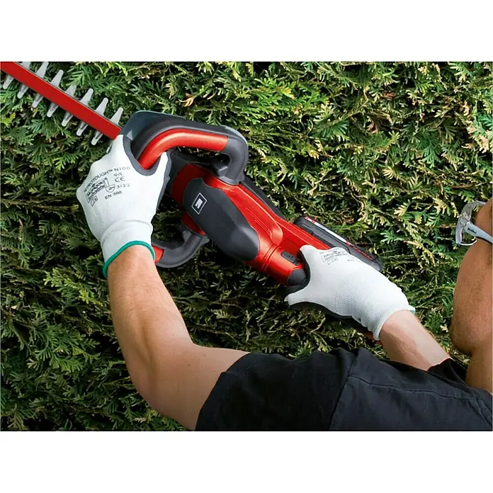 Einhell Power X-Change 18V Akku-Heckenschere GC-CH 1846 Li-Solo - 18 V, Ohne Akku, Schnittlänge: 46 cm - Eine Person bedient eine rote Heckenschere vor einer grünen Hecke, geschützt durch weiße Handschuhe für sicheres Arbeiten.