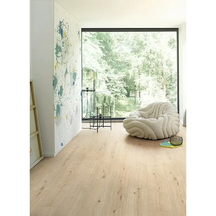 Parador Designboden Modular One - Eiche Atmosphere geschliffen, Landhausdiele, 1.285 x 194 x 8 mm - Helles Holzlaminat erhellt einen modernen Raum mit Kunst, einer Lampe und einem bequemen Sessel vor einem Fenster mit Blick auf üppiges Grün.