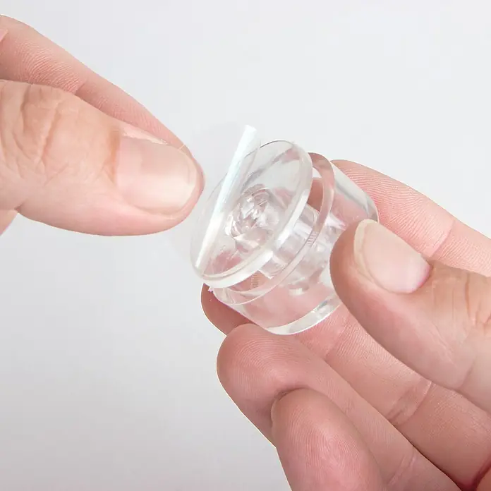 Walteco Türstopper - Kunststoff, B x L: 33 x 100 mm - Nahaufnahme eines transparenten Möbelgleiters mit Klebefläche, der von zwei Händen gehalten wird. Ideal zum Schutz von Böden.