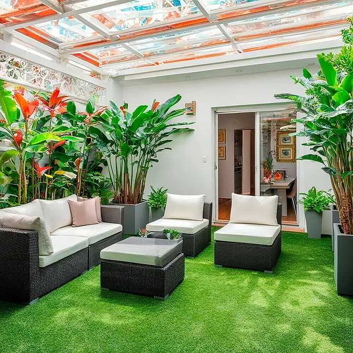 Césped artificial Dresde - 5 x 2 m, Altura del pelo: 30 mm, Densidad: 19.000 puntadas/m² - Una idílica escena de jardín con muebles de ratán, vegetación exuberante y un techo de cristal luminoso y soleado para relajarse al aire libre.