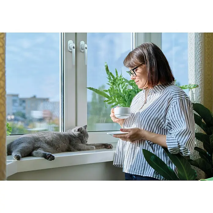 Basi Fenstergriff FG 503 - Weiß, Distanznorm: 43 mm - Frau mit einer Tasse Kaffee am Fenster, neben einer entspannten Katze. Gemütliche Szene mit Zimmerpflanzen und Tageslicht.