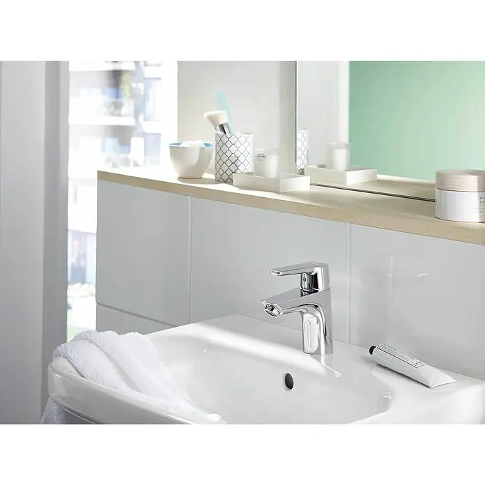 Hansa Polo Waschtischarmatur S-Size - Chrom, Glänzend - Ein glänzender Chrom-Wasserhahn auf einem weißen Waschbecken, ergänzt durch eine Tube Handcreme und ein flauschiges weißes Handtuch für ein sauberes Ambiente.