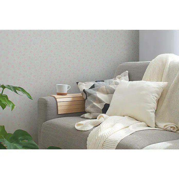 A.S. Création Maison Charme Vliestapete Blümchen - Grau-Rosa-Weiß, Floral, 10,05 x 0,53 m - Eine stilvolle Wohnzimmerszene mit einem grauen Sofa, dekoriert mit Kissen und einer Strickdecke, neben einer Zimmerpflanze und einer Tasse.