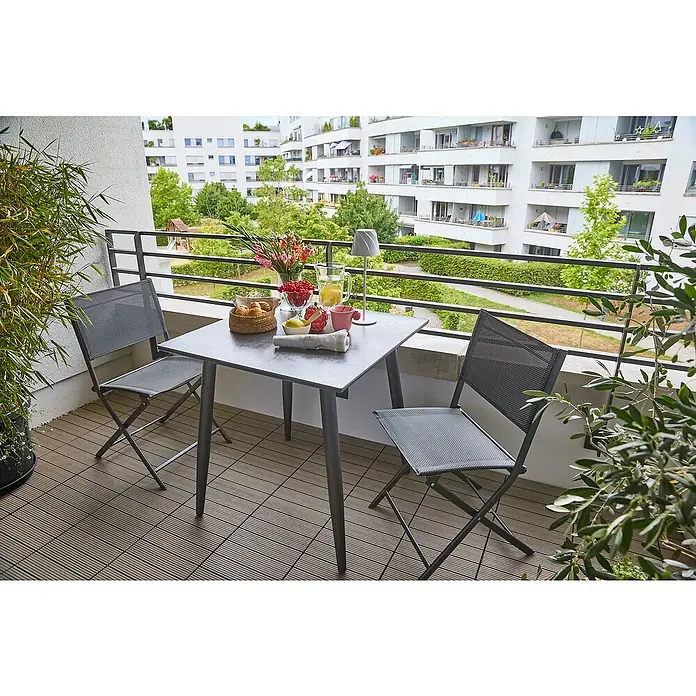 Sunfun Balkonmöbel-Set Solo/Maja - 3 -tlg., Aluminium, Sintered Stone, Textilen, Night-Grey, Gartenstuhl klappbar - Balkonansicht mit grauem Gartentisch-Set, inklusive Tisch und zwei Stühlen, gedeckt mit Frühstücksleckereien und Blumen, vor dem Hintergrund moderner Wohnhäuser.