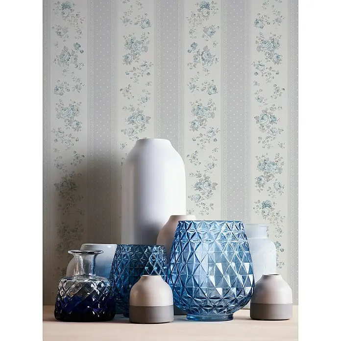 A.S. Création Maison Charme Vliestapete Vertikale Blümchen - Grau-Blau-Weiß, Floral, 10,05 x 0,53 m - Eine Ansammlung dekorativer Vasen in verschiedenen Formen und Farben, darunter blaue Glasvasen und moderne Keramikvasen, vor einer floralen Tapete.