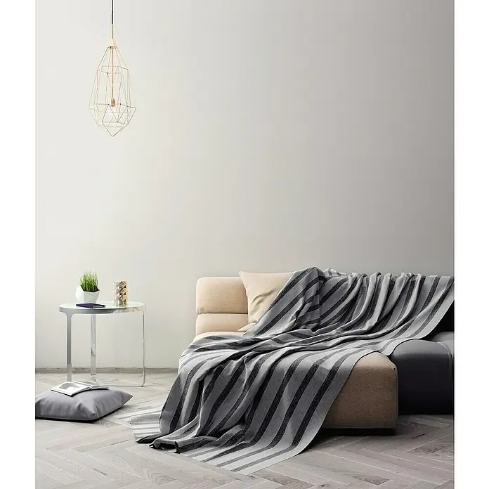 A.S. Création Maison Charme Vliestapete - Creme-Hellgrau, Uni, 10,05 x 0,53 m - Eine gemütliche Couch mit einer gestreiften Tagesdecke in einem hellen Raum. Die Lampe und der Beistelltisch runden das moderne Ambiente ab.