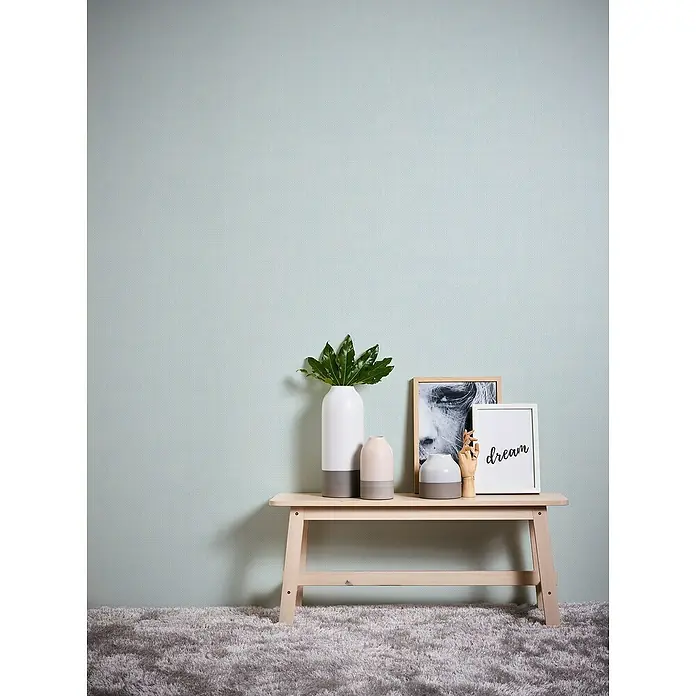 A.S. Création Maison Charme Vliestapete Kleinmuster - Blau-Weiß, Uni, 10,05 x 0,53 m - Dekorative Inneneinrichtung mit einem hellen Holztisch, Vasen, Kunstobjekten und einem gerahmten "Dream"-Schriftzug vor einer hellblauen Wand.