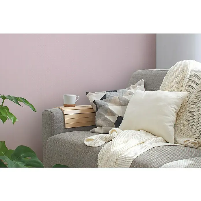 A.S. Création Maison Charme Vliestapete Punkte - Rosa-Weiß, Grafisch, 10,05 x 0,53 m - Nahaufnahme eines grauen Sofas mit Kissen und einer Strickdecke, ergänzt durch ein Holztablett mit einer Tasse, vor einem rosa Hintergrund.
