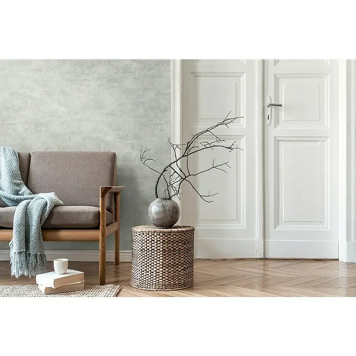 A.S. Création Daniel Hechter 7 Vliestapete Putz - Graublau, Steinoptik, 10,05 x 0,53 m - Modernes Wohnzimmer Interieur mit einem bequemen Sessel, einem Beistelltisch mit Dekoration und einer hellen Wand für eine ruhige Atmosphäre.