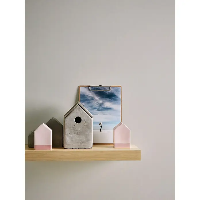 A.S. Création Maison Charme Vliestapete - Creme-Grau, Uni, 10,05 x 0,53 m - Dekorative Miniaturhäuser auf einem Holzregal, einige mit rosa Akzenten, vor einem Clipboard mit einer Himmelsaufnahme.