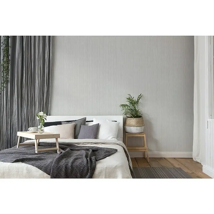 A.S. Création Maison Charme Vliestapete - Grau-Hellgrau, Streifen, 10,05 x 0,53 m - Zimmer mit einem bequemen Bett, das mit Kissen und einer Decke dekoriert ist, und einem Holzhocker mit einer Topfpflanze.