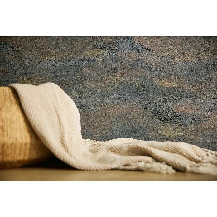 A.S. Création Metropolitan Stories Hot Spots Vliestapete Wellengrafik - Blau-Grau-Metallic, Wellen, 10,05 x 0,53 m - Nahaufnahme eines beigen Sofas mit Fischgrätenmuster und Fransen auf einem Holztisch vor einem gemusterten Hintergrund.
