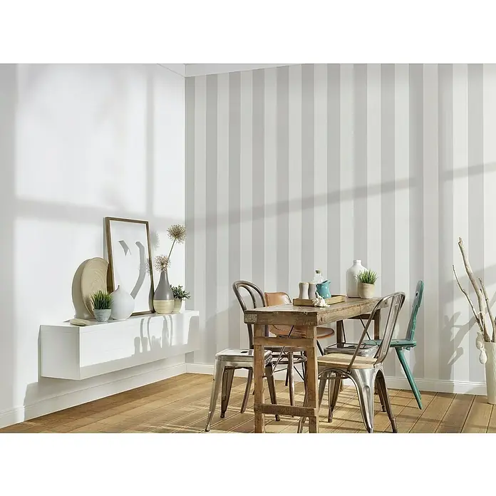 A.S. Création Maison Charme Vliestapete Blockstreifen - Grau-Creme-Weiß, Streifen, 10,05 x 0,53 m - Ein Esstisch mit verschiedenen Stühlen und dekorativen Elementen vor einer gestreiften Wand. Der Tisch ist das zentrale Element des Bildes.