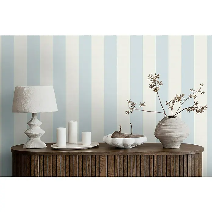 A.S. Création Maison Charme Vliestapete Blockstreifen - Blau-Weiß, Streifen, 10,05 x 0,53 m - Stilvolles Interieur mit einer Lampe, Kerzen, einer Schale mit Birnen und einer Vase mit Zweigen auf einem Holzschrank vor gestreifter Wand.