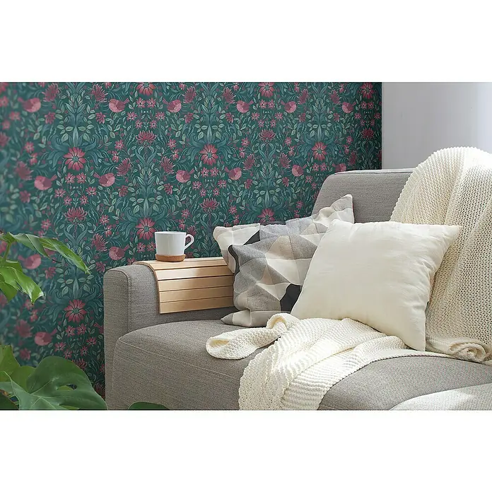 A.S. Création Maison Charme Vliestapete Blumen - Dunkelgrün-Pink-Petrol, Floral, 10,05 x 0,53 m - Einrichtungsidee mit einem grauen Sofa, Zierkissen und einer Strickdecke vor einer floralen Tapete mit Vögeln.