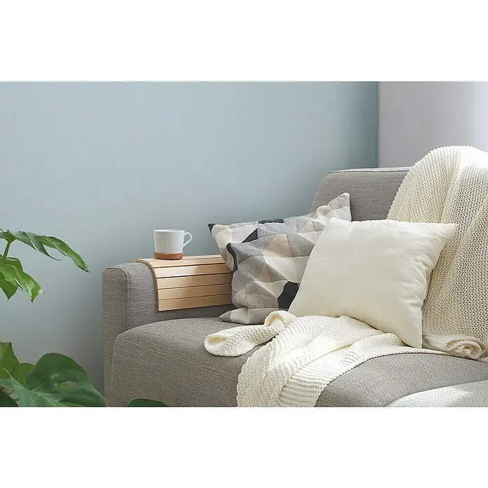A.S. Création Maison Charme Vliestapete - Blau-Hellblau, Uni, 10,05 x 0,53 m - Nahaufnahme eines grauen Sofas mit Kissen, einer Strickdecke und einer Holzablage mit einer weißen Tasse darauf.