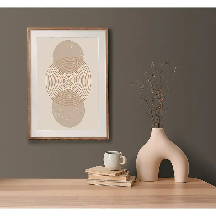 A.S. Création Pintwalls 2 Vliestapete - Braun, Uni, 10,05 x 0,53 m - Ein gerahmtes Kunstwerk mit abstrakten Kreismotiven hängt an einer Wand über einem Tisch mit Dekorationen wie Büchern und einer Vase.