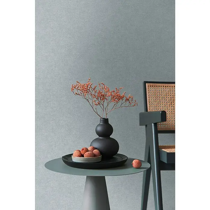 A.S. Création Pintwalls 2 Vliestapete - Grau-Blau, Uni, 10,05 x 0,53 m - Nahaufnahme eines Tisches mit einer schwarzen Vase und einer Schale mit roten Früchten, neben einem Stuhl.