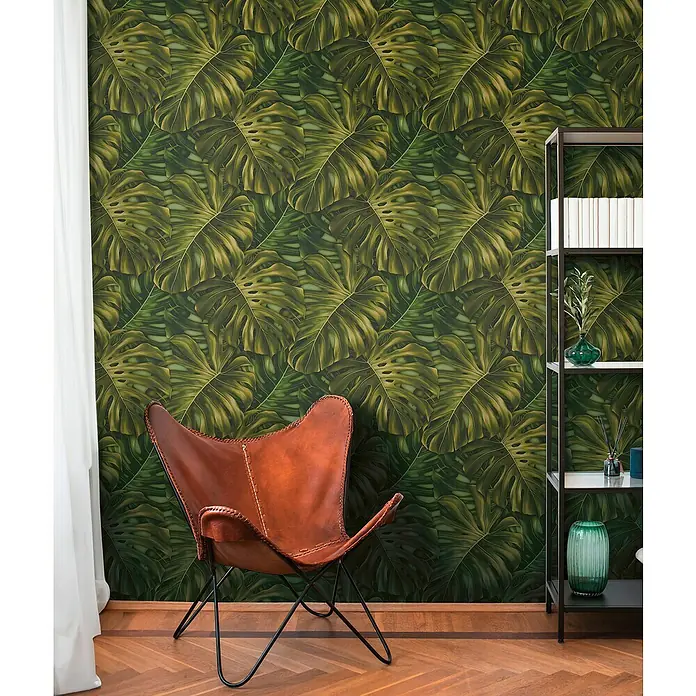A.S. Création Pintwalls 2 Vliestapete Dschungel - Grün-Schwarz-Braun, Floral, 10,05 x 0,53 m - Lederner Butterfly-Stuhl vor einer Wand mit tropischem Monstera-Blatt-Muster, neben einem Regal mit Dekorationsgegenständen.