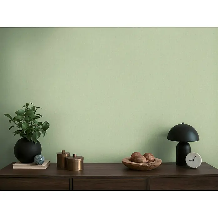 A.S. Création Michalsky Metropolis Vliestapete - Grün, Uni, 10,05 x 0,53 m - Nahaufnahme eines stilvollen Sideboards mit Dekorationen wie einer Lampe, einer Pflanze und dekorativen Accessoires auf einer hellgrünen Wand.