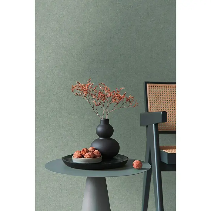 A.S. Création Metropolitan Stories Hot Spots Vliestapete - Grün, Uni, 10,05 x 0,53 m - Elegantes Stillleben mit einer schwarzen Vase, Trockenblumen und Litschis auf einem grauen Tisch, ergänzt durch einen stilvollen Stuhl im Hintergrund.