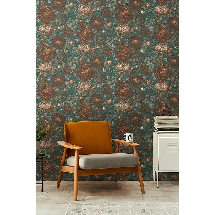 A.S. Création Michalsky Metropolis Vliestapete Blüten - Grün-Grau-Orange, Floral, 10,05 x 0,53 m - Das Foto zeigt eine Wand mit einer floralen Tapete in dunklen Farben und einen bequemen Sessel mit hellgrauer Sitzfläche.
