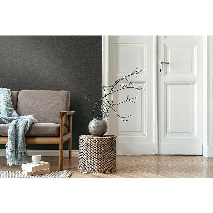 A.S. Création Daniel Hechter 7 Vliestapete - Schwarz, Betonoptik, 10,05 x 0,53 m - Das Bild zeigt ein stilvolles Wohnzimmer mit einem grauen Sofa, einem runden Tisch mit einer Vase und einem Bücherstapel mit einer Tasse darauf.