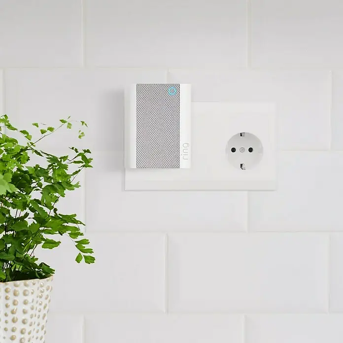 Ring Set Türklingel mit Kamera Video Doorbell Akku Plus + Funk-Gong Chime 2. Gen. - Nickel, 1.920 x 1.080 Pixel (Full HD), 2,8 x 6,2 x 12,8 cm - Ring-Gerät an einer weißen Wand neben einer Steckdose. Das Gerät ist weiß mit einem grauen Gitter und einem blauen Ring.