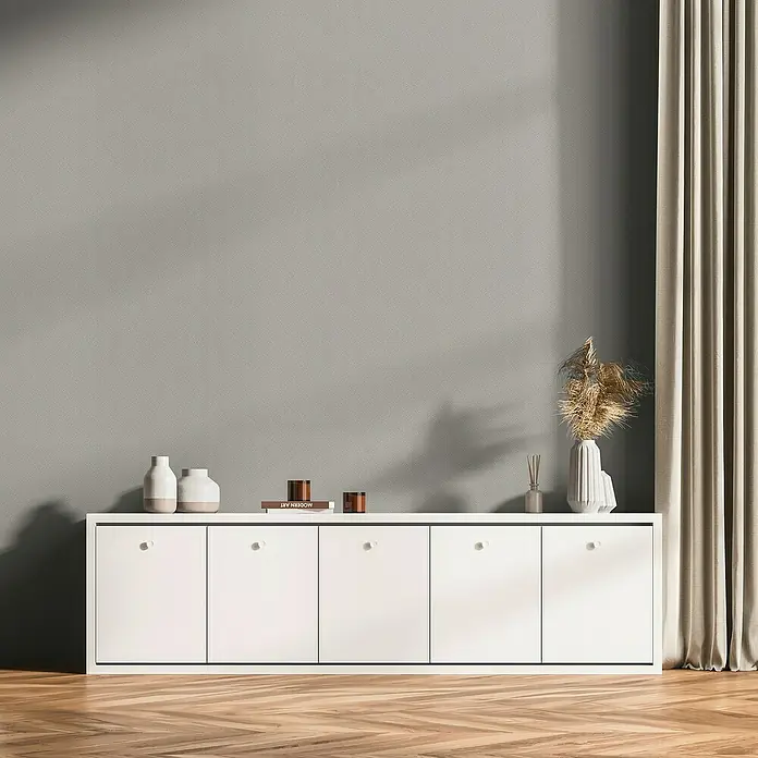 A.S. Création Daniel Hechter 7 Vliestapete 3D-Grafikstreifen - Hellgrau-Metallic, Grafisch, 10,05 x 0,53 m - Ein weißes Sideboard mit dekorativen Elementen wie Vasen, Kerzen und einem Trockenblumenstrauß vor einer grauen Wand.