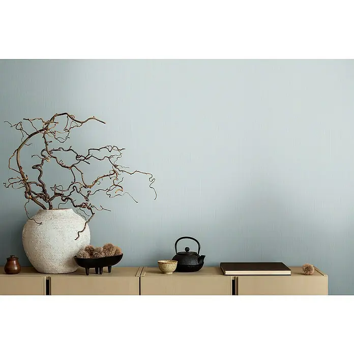 A.S. Création Michalsky Metropolis Vliestapete - Pastellblau, Uni, 10,05 x 0,53 m - Stillleben mit Keramikvase, Teekanne und Buch auf einem beigen Sideboard vor hellblauem Hintergrund.