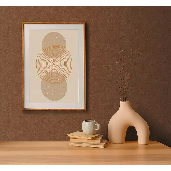 A.S. Création Pintwalls 2 Vliestapete - Rot-Braun, Uni, 10,05 x 0,53 m - Ein gerahmtes Kunstwerk mit Kreismuster hängt über einem hellen Holztisch mit Dekorationen wie Bücher, Tasse und einer Vase mit Zweigen.