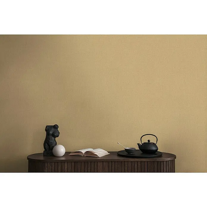 A.S. Création Michalsky Metropolis Vliestapete - Beige-Gold, Uni, 10,05 x 0,53 m - Nahaufnahme eines eleganten Holztisches mit Skulptur, offenem Buch und Tee-Set vor einem ockerfarbenen Hintergrund.
