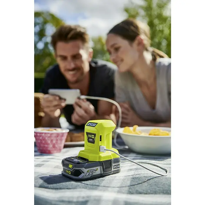 Ryobi ONE+ Adaptador USB R18USB - 18 V, Sin batería - Esta imagen muestra un adaptador de batería Ryobi ONE+ con un puerto USB, que alimenta un smartphone al aire libre.