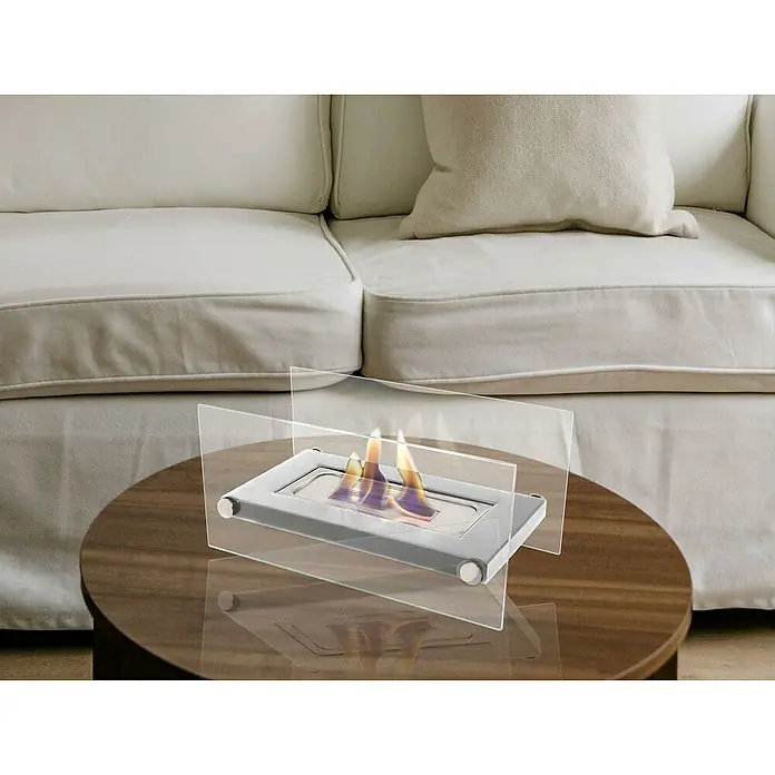 Purline Chimenea de bioetanol Oniros - Acero inoxidable, Plateado - Una chimenea de mesa con cerramiento de vidrio se asienta sobre una mesa redonda de madera frente a un sofá beige, añadiendo calidez.