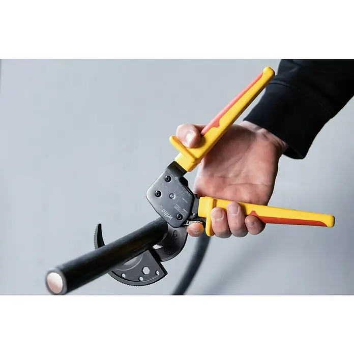 Wiha Ratschenkabelschneider VDE 300 MM² - Länge: 27,5 cm - Eine Person hält eine Wiha Kabelschere mit gelb-roten Griffen, die ein schwarzes Kabel schneidet.