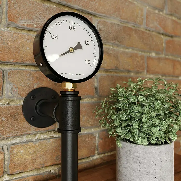 Walteco Rusticline Manometer - Stahl, Schwarz - Nahaufnahme eines schwarzen Manometers mit Ziffernblatt und Zeiger, befestigt an einer Wand mit Rohren.
