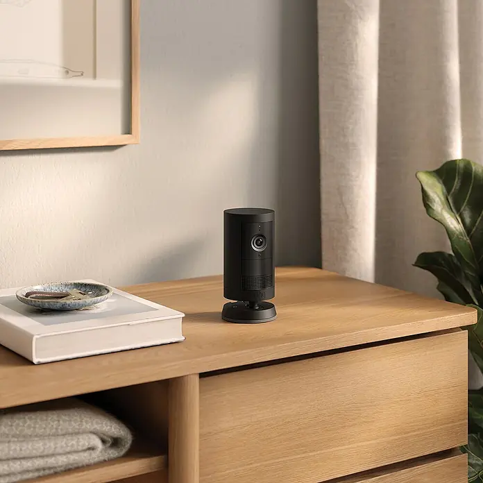 Ring Außenkamera Akku Plus - 2.048 x 1.536 Pixel (2K-HD), Schwarz - Das Ring Sicherheitskamera-Stick Up Cam Essential, präsentiert auf einem Tisch mit dekorativen Elementen, bietet Sicherheit und Stil für Ihr Zuhause.
