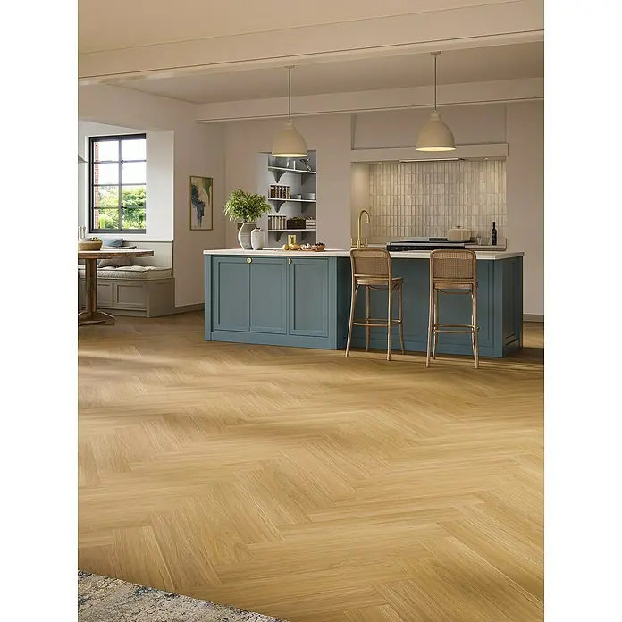 Parador Vinylboden Trendtime 3 - Eiche Oxford karamellbraun, Fischgrät, 730 x 146 x 5 mm - Helles Wohnkücheninterieur mit Fischgrätparkettboden, einer blauen Kücheninsel und zwei Barhockern unter Pendelleuchten.
