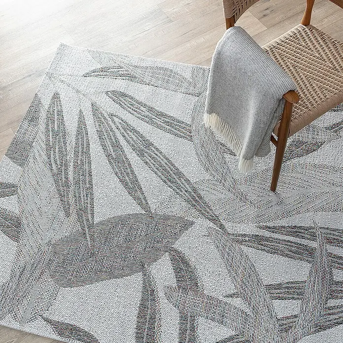 Dekoria Flachgewebeteppich - beige, braun, bunt, 230 x 160 cm - Ein grauer Teppich mit Blattmuster liegt auf einem hellen Holzboden, teilweise von einem Stuhl mit einer hellgrauen Decke bedeckt.