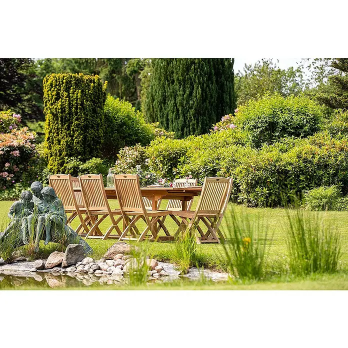 Kai Wiechmann Gartenstuhl - Teakholz, Klappbar, Naturbelassen, B x T x H: 47 x 60 x 94 cm - Ein einladender Gartentisch mit Stühlen aus Holz auf einem grünen Rasen, umgeben von üppigem Grün und einem ruhigen Teich mit Steinskulptur.