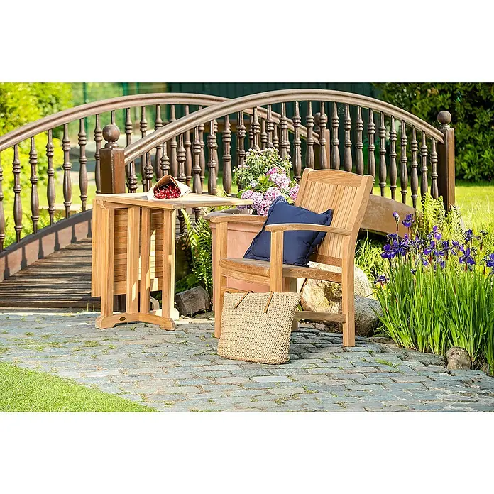 Kai Wiechmann Gartensessel - Teakholz, Naturbelassen, B x T x H: 60 x 65 x 92 cm - Ein Gartenset mit einem Holztisch, einem Stuhl mit blauem Kissen und einer Korbtasche vor einer dekorativen Brücke, idyllisch im Garten.