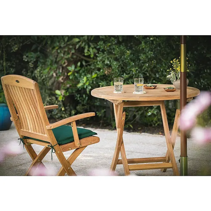 Kai Wiechmann Sitzauflage - Klappstuhl, L x B x H: 35 x 40 x 4 cm, Polyacryl, Marineblau - Ein Teakholz-Gartenset mit einem runden Tisch, zwei Gläsern und einem Stuhl mit grüner Polsterung vor einem grünen Hintergrund.