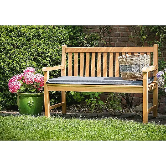 Kai Wiechmann Gartenbank - B x T x H: 150 x 65 x 92 cm, Teak, Naturbelassen - Eine Gartenbank aus Holz mit einem grauen Kissen und einem Korb darauf, umgeben von Pflanzen und Grünflächen.