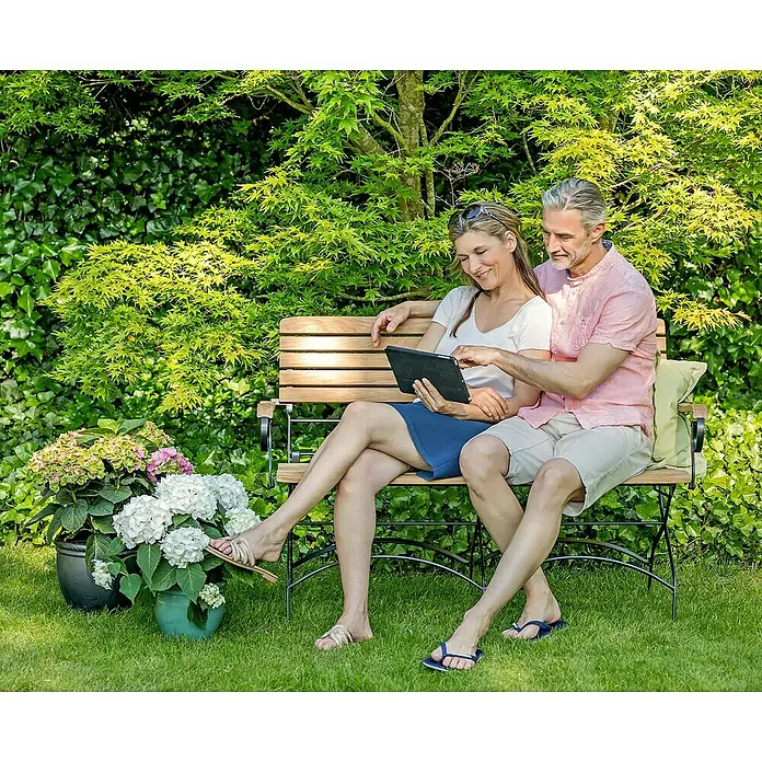 Kai Wiechmann Gartenbank - B x T x H: 131 x 59 x 88 cm, Teak, Naturbelassen, Klappbar - Ein Paar sitzt auf einer Gartenbank und betrachtet ein Tablet, umgeben von üppigem Grün. Die Bank bietet eine gemütliche Sitzgelegenheit im Freien.